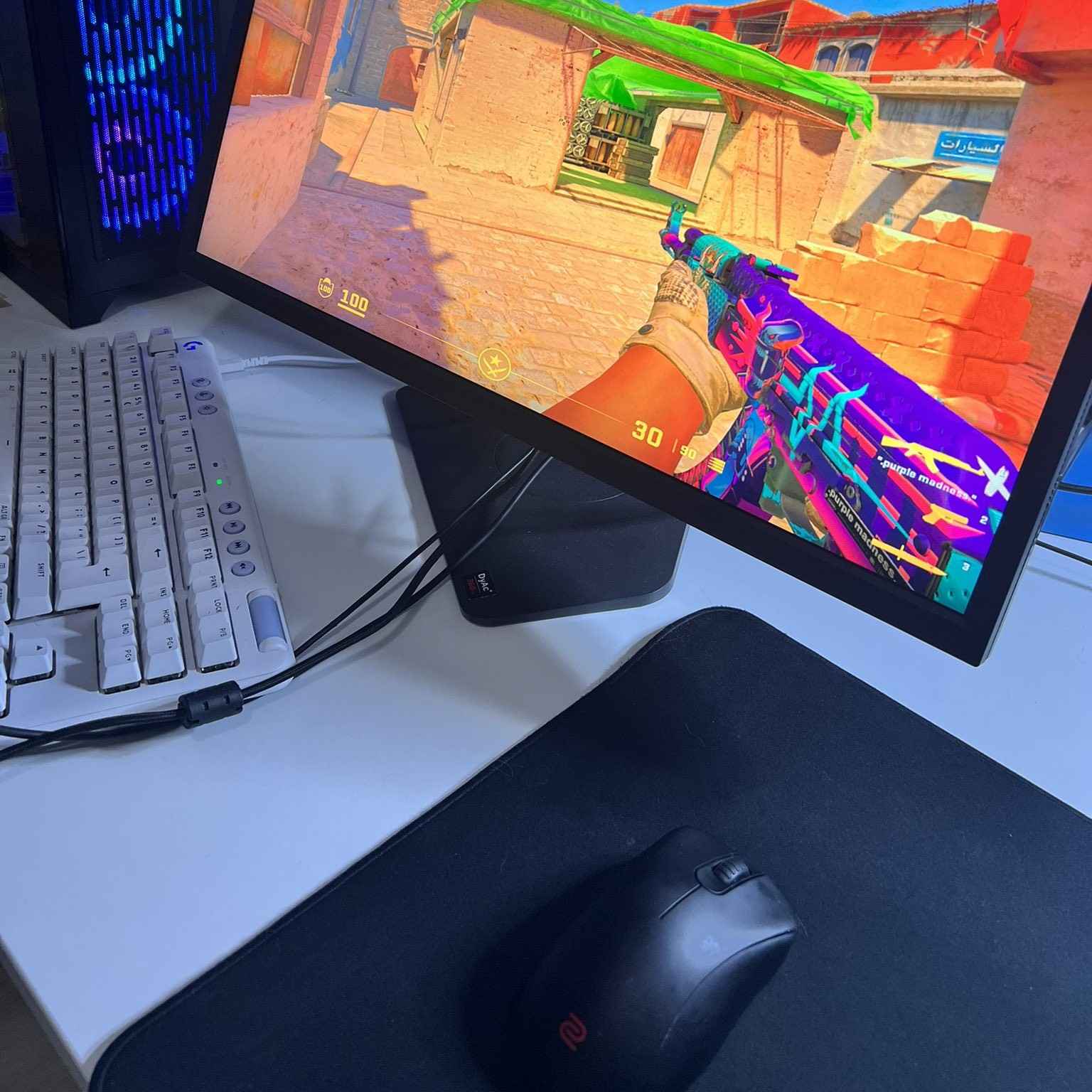 Gaming setup with a monitor displaying Counter-Strike 2 gameplay, a white keyboard, and a black mouse on a mousepad.