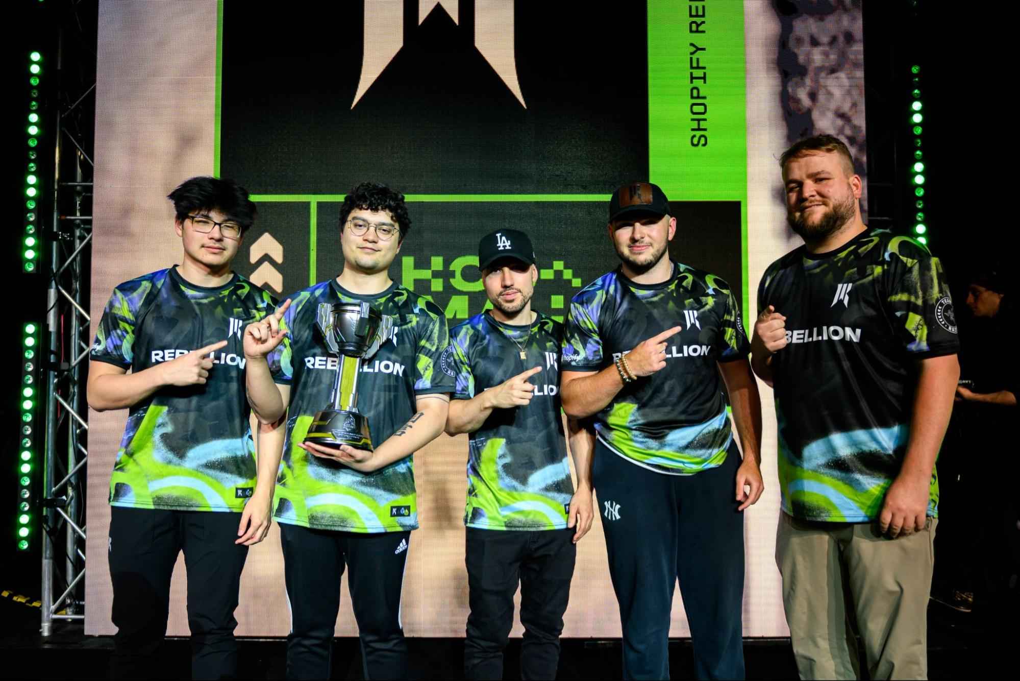 Five esports players in colorful jerseys pose on stage, holding a trophy, celebrating their victory against a vibrant backdrop.