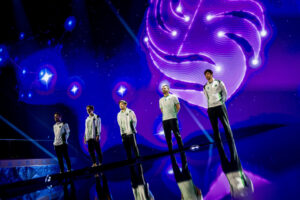 Five individuals stand in line, wearing matching white jackets, on a stage with a shiny floor. A cosmic backdrop of swirling purple and blue lights creates an ethereal atmosphere