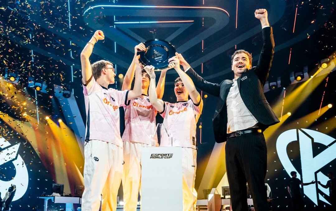 A team of esports players celebrates their victory, holding a trophy amidst confetti during the Esports World Cup, with vibrant stage lighting.