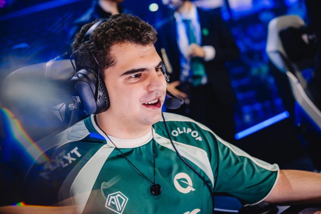 Esports player in a green team jersey wearing a headset and smiling during a match.