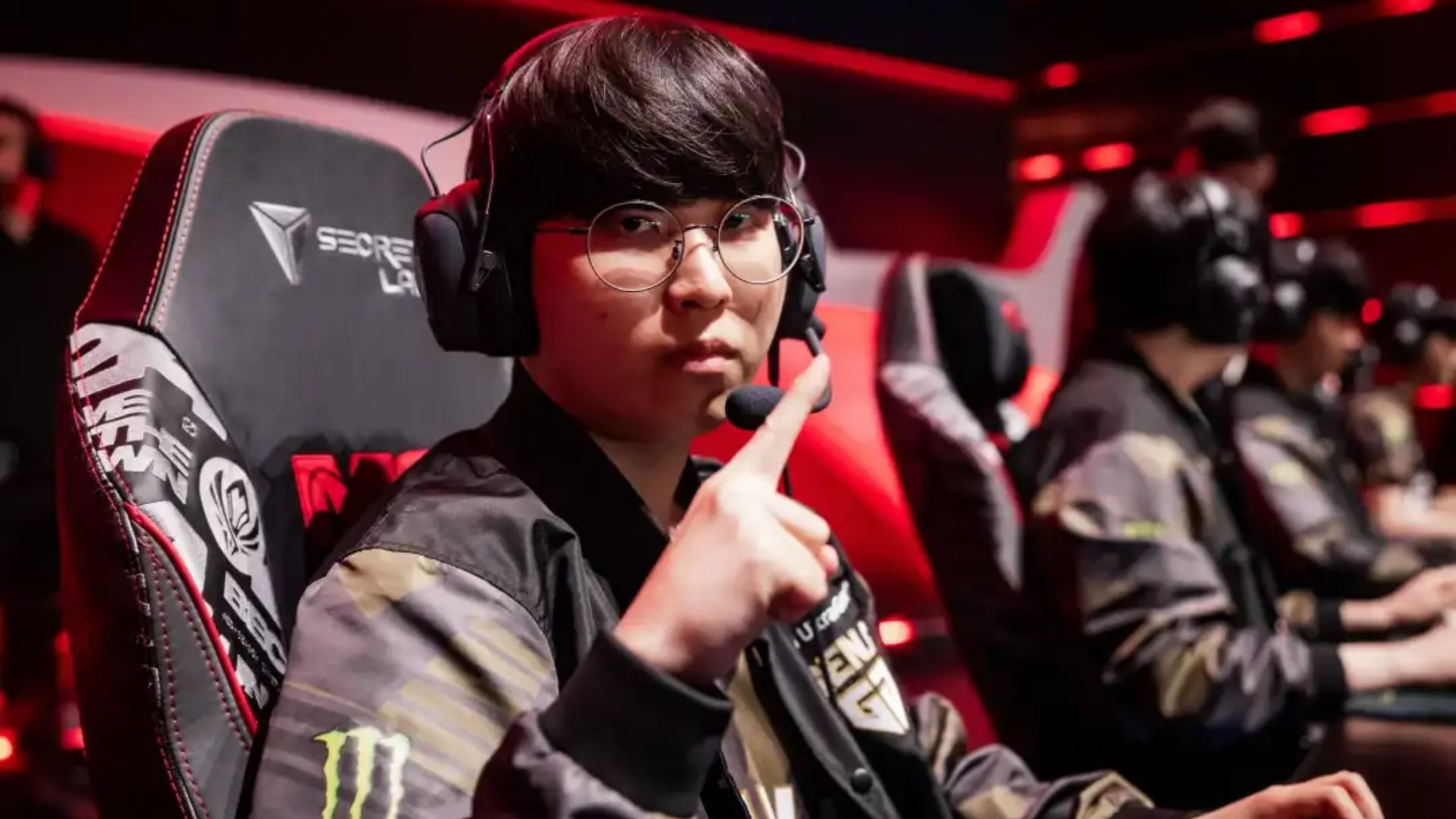 Gen.G Kiin sitting at his gaming setup in team jacket and headset, gesturing toward the camera