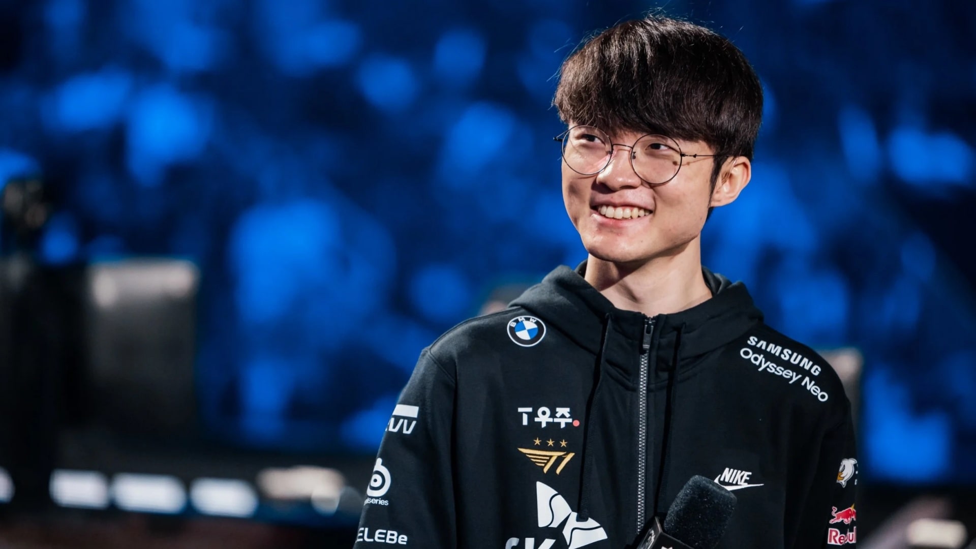 T1 Faker smiling on stage, wearing a team jacket and holding a microphone