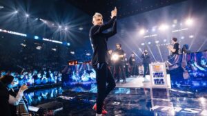 A League of Legends event featuring players and casters on stage under bright arena lights