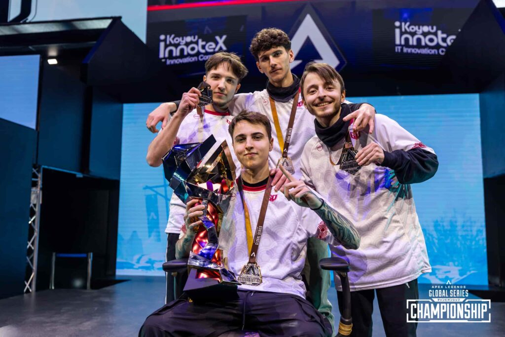 A team of four Apex Legends players celebrating on stage with the ALGS Championship trophy, smiling and holding their medals after winning the event