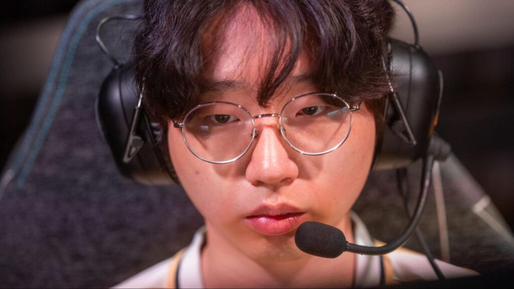 A professional esports player wearing a headset and blue team jersey smiles while competing in a live gaming event.