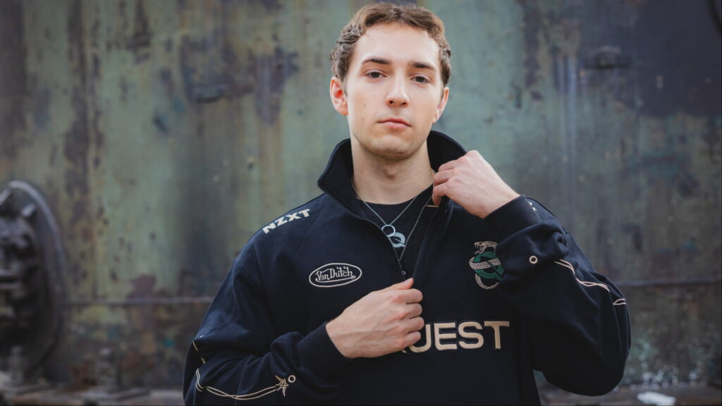 A man in a black esports jacket adjusts his collar against a textured metal backdrop