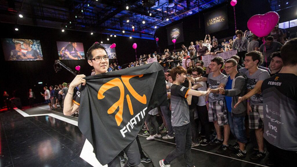 A FNATIC player proudly holds the team’s flag while greeting fans after a League of Legends match, with a cheering crowd and bright stage lights in the background.