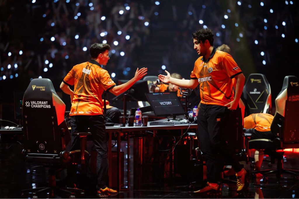 FNATIC players in orange jerseys celebrating a win at Valorant Champions, sharing a high-five on stage surrounded by glowing audience lights and gaming setups.