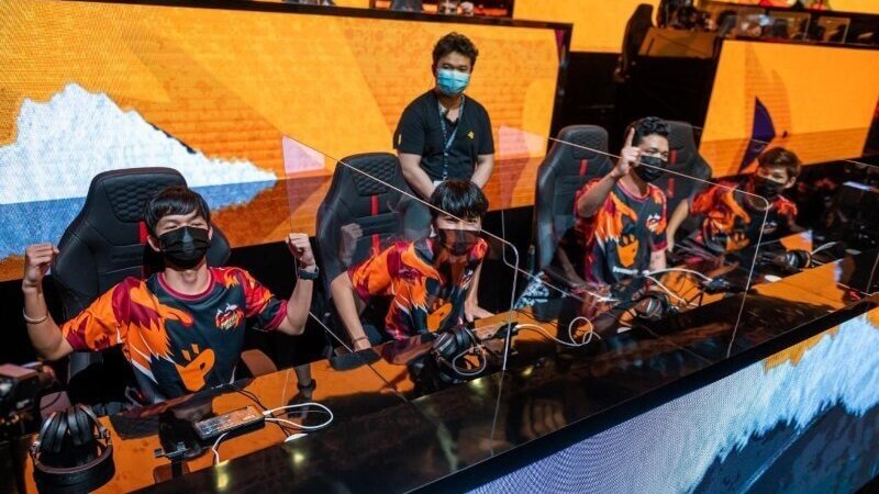 The winning team celebrating at the Free Fire World Series 2021, sitting at their gaming stations in orange and black jerseys with clear dividers and headsets ready for competition.
