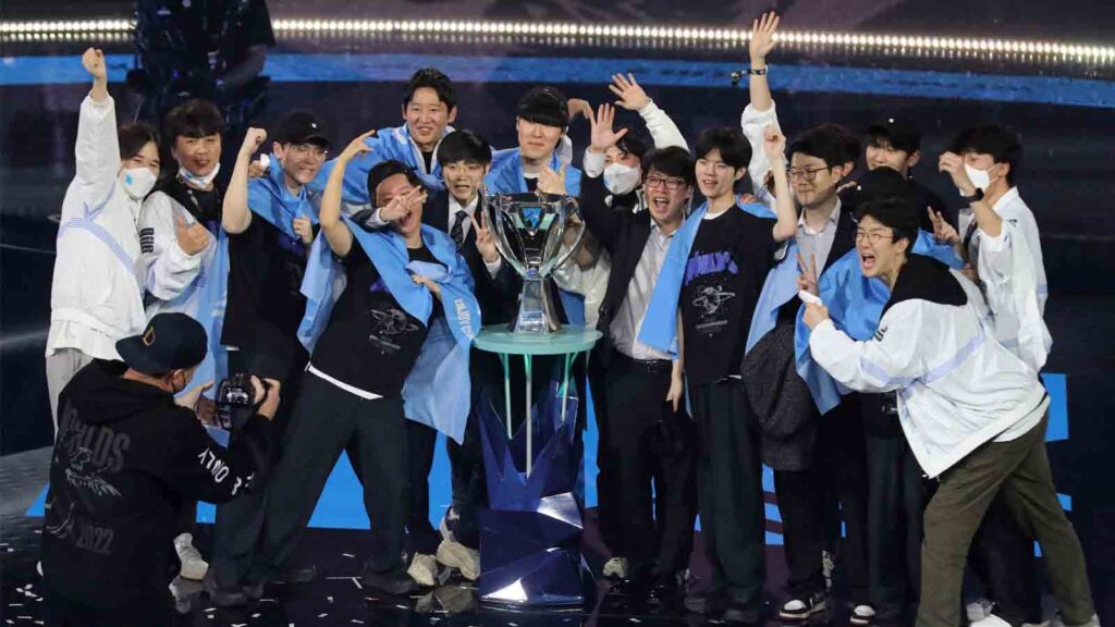 The League of Legends World Championship 2022 winning team celebrating around the Summoner’s Cup, cheering and posing with blue team scarves under the stage lights.