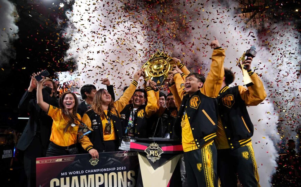 The M5 World Championship 2023 winners celebrating on stage with confetti and smoke effects, holding the golden championship trophy while wearing black and yellow team jackets.