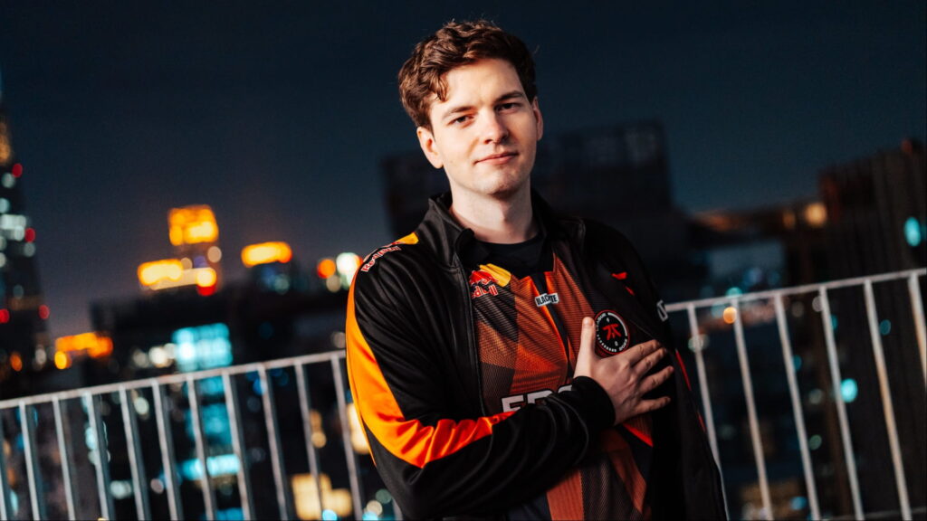 A man in an orange-black esports jersey stands on a balcony with city lights behind him