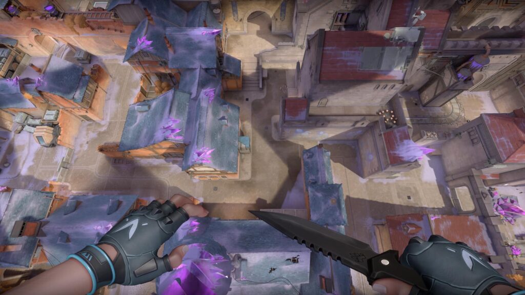 A player in VALORANT looks down from a high ledge at snowy rooftops covered in purple crystals. The view shows a quiet street below as the agent prepares to jump with a knife in hand