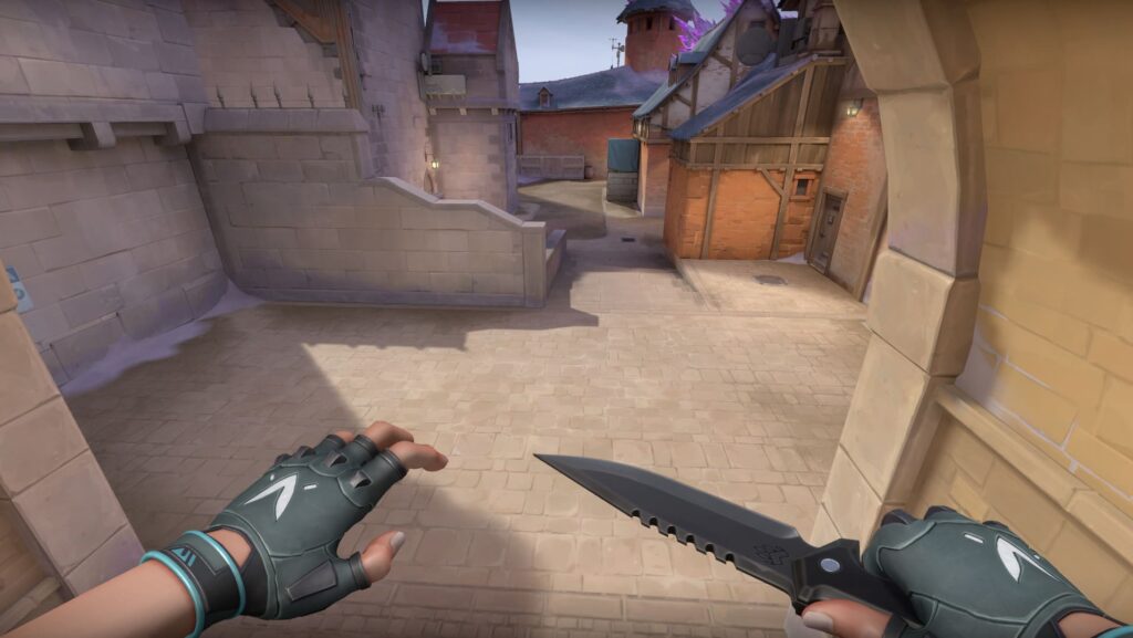 A VALORANT agent moves toward a quiet alley with a knife ready. The sun casts soft shadows across the stone path and wooden buildings