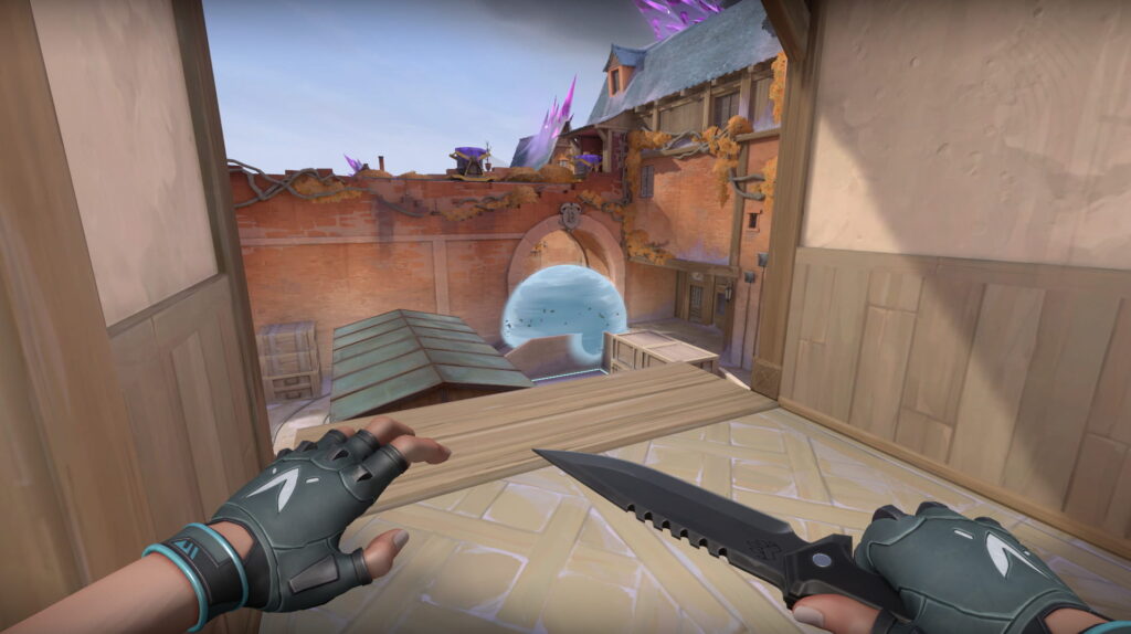 A VALORANT agent watches a blue smoke cloud form under the archway. Crates and rooftops frame the entry as the player holds a knife