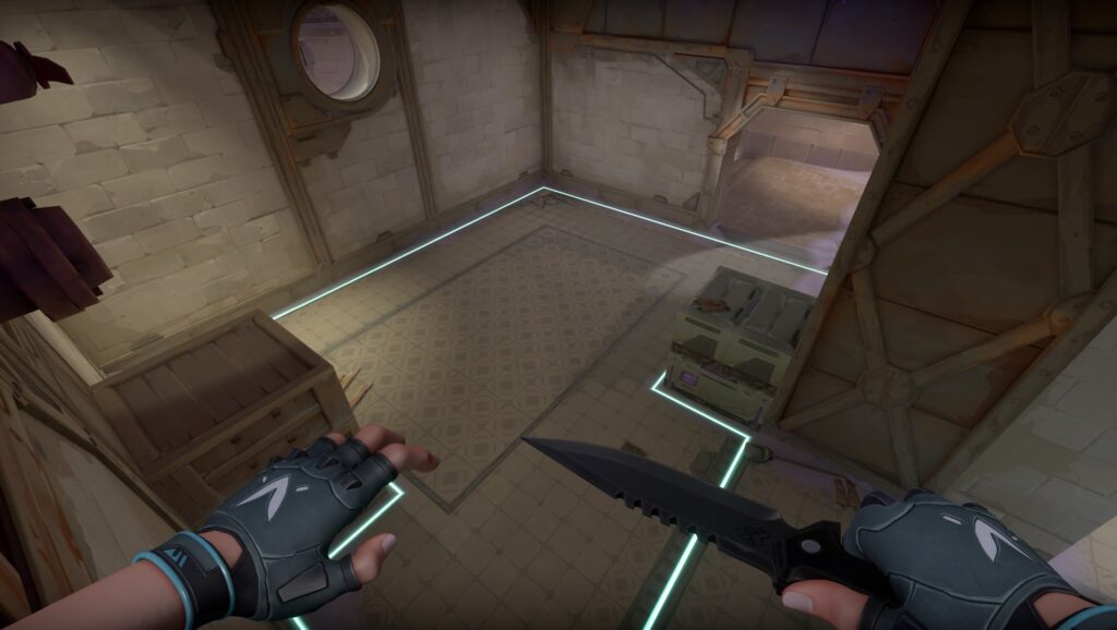 A VALORANT agent looks down into a small marked site room with crates and glowing boundary lines. The space is quiet with pale walls and metal supports