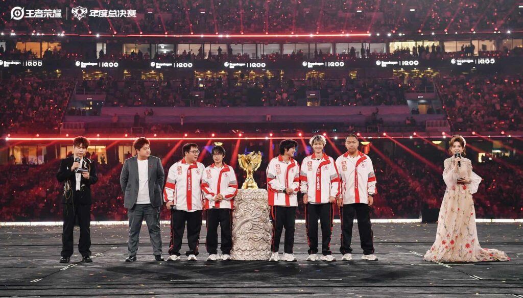 A team of Honor of Kings esports players stands on stage around a golden trophy during a large arena championship ceremony, with presenters on both sides and a cheering crowd in the background