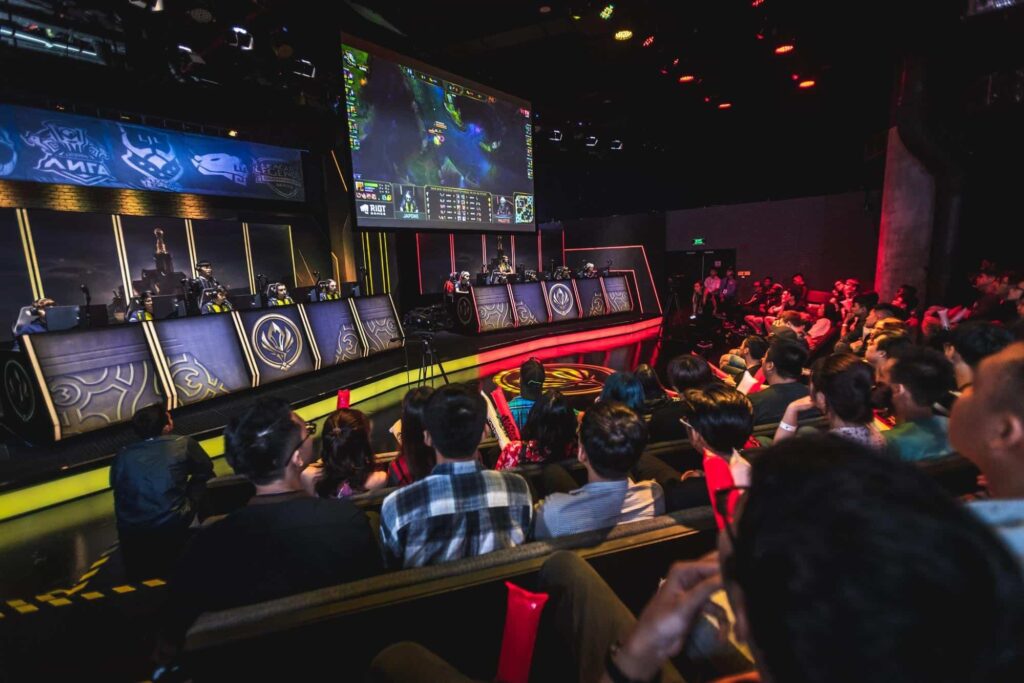 A large crowd watches a live League of Legends esports match in an arena, with two teams competing on stage beneath a big screen showing in-game action