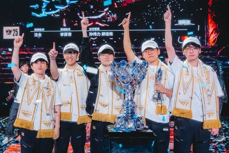 A team of five esports players celebrating on stage with a large silver championship trophy, wearing matching white outfits and pointing upward as confetti falls around them