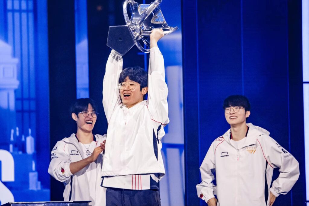 Three esports players in white team jackets celebrate on stage as the center player lifts a large silver trophy overhead, smiling under bright blue stage lighting