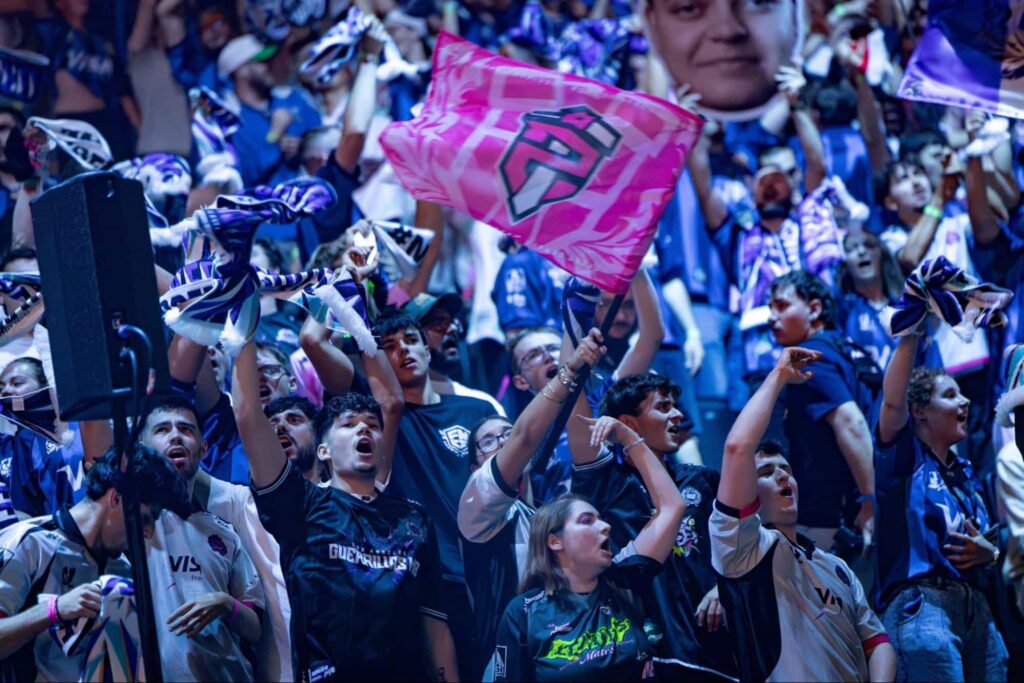 Esports fans wave team flags and cheer loudly inside a packed tournament arena