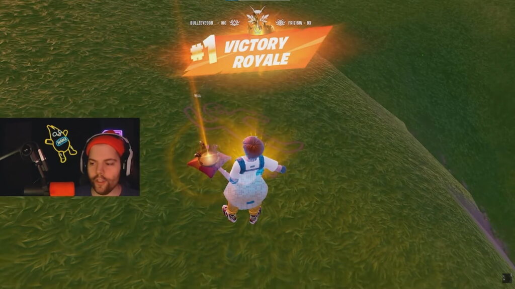 Fortnite player celebrates a #1 Victory Royale on a grassy hill, with a live streamer facecam visible in the corner during the winning moment