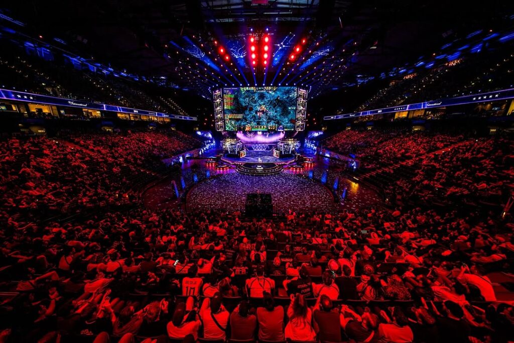 A packed arena hosting a live Dota 2 esports tournament with a large stage and in-game action on the main screen