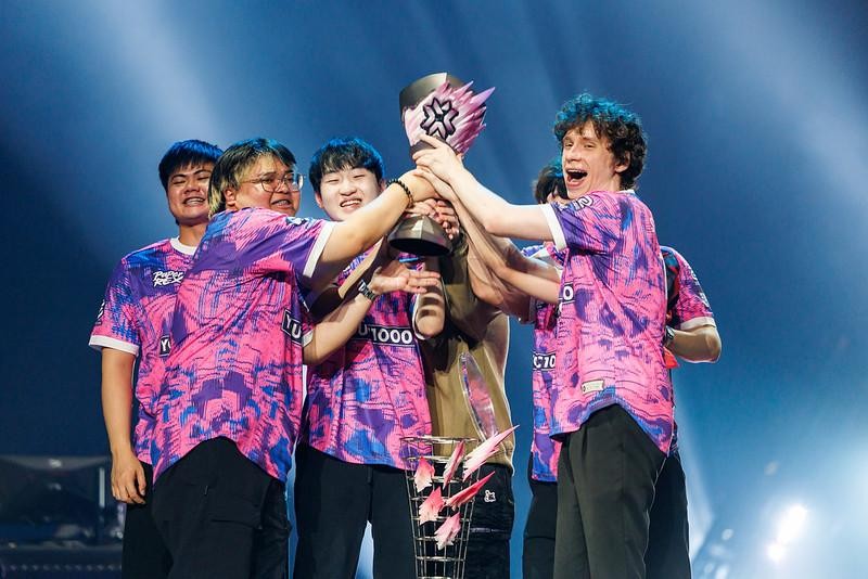 A group of six people in matching pink and purple jerseys celebrating with a trophy on stage