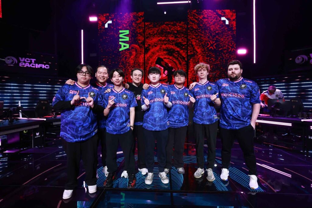 FNATIC VALORANT players posing together on stage after a victory, wearing black and orange jerseys at a major VCT EMEA event, led by Jake “Boaster” Howlett and Emil “Alfajer” Ali Beder.